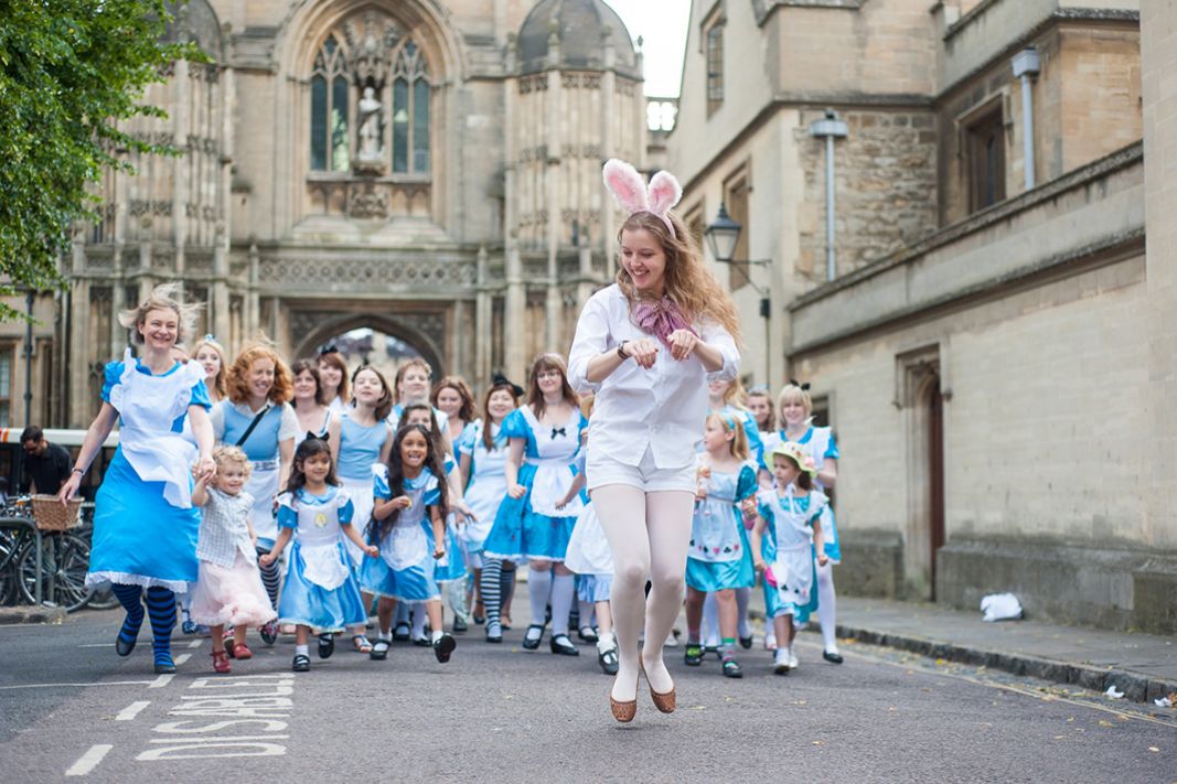 A woman dressed as the White Rabbit jumping with lots of girls dressed as Alice following