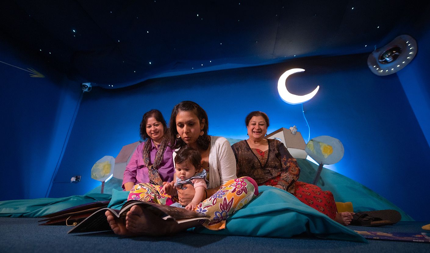 A baby and family members sat in a dark cosy space with a light up moon readying a book.