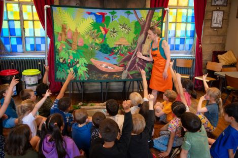 A group of children with their hands up in front of a Story Leader telling a story